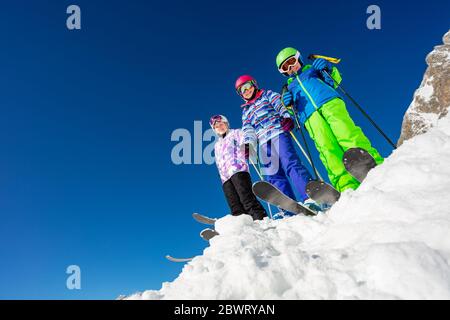 Vista dal basso di un gruppo di tre bambini in piedi sulla cima della montagna con neve indossando sci colorato vestito sopra il cielo blu Foto Stock