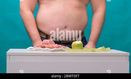 l'uomo con una pancia grassa sceglie tra salsiccia e mela verde. Il concetto di dieta e di volontà. Foto Stock