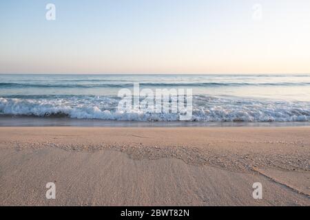 Due linee orizzontali dividono il cielo dalla terra, sabbia dorata con conchiglie e calme onde di mare mentre il sole scende. Foto Stock