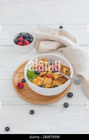 Prima colazione ciotola bianca con minuscoli cereali pancake con mirtilli, lamponi e menta su sfondo di legno Foto Stock