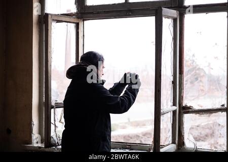 Uomo adulto con cappotto invernale freddo nero che scatta foto della vista esterna dalla finestra rotta di una casa abbondata in una fredda giornata invernale. Foto Stock