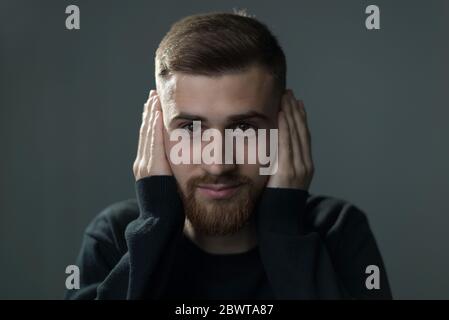 ritratto di un ragazzo giovane, sorridente, copre le orecchie con le mani, guarda la fotocamera. Foto di studio su sfondo grigio. Non è vero, mi sta Foto Stock