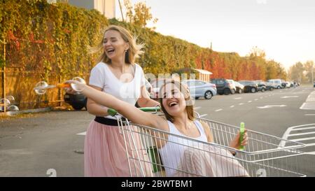 Ritratto di due ragazze ridenti che soffiano bolle di sapone mentre si cavalcano nel carrello per lo shopping sul parcheggio dell'auto presso il grande centro commerciale Foto Stock