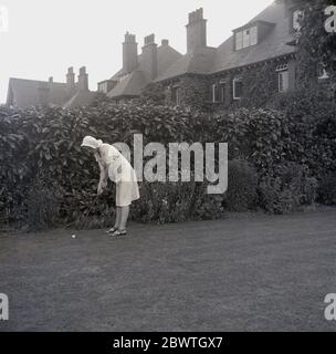 Anni 60, storico, in piedi accanto ad una siepe alta sul prato all'aperto in un giardino nel suburban Inghilterra, una giovane donna che indossa un cofano e dresss tenendo un putter tradizionale forma in piedi in piedi pronto a putt una palla da golf lungo l'erba. Foto Stock