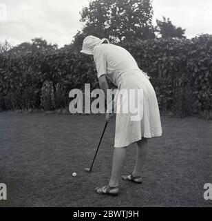 1960, storico, in piedi su un prato in un giardino di backgarden nella periferia dell'Inghilterra, una giovane donna che indossa un cofano e vestito con un putter tradizionale a forma di in piedi in piedi pronti a putt una palla da golf lungo l'erba. Foto Stock