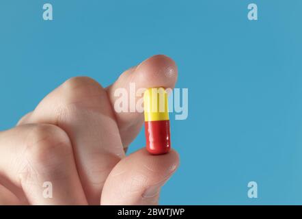 Dita che tengono in su una capsula di medicinale gialla e rossa. Closeup macro estremo con sfondo blu Foto Stock