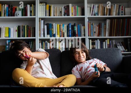 Fratelli che giocano un videogioco mentre si siedono sul divano contro la libreria nel soggiorno a casa Foto Stock