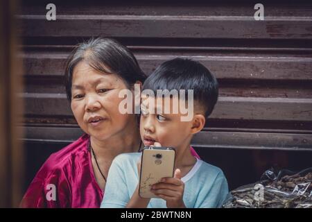 Feng Huang, Cina - Agosto 2019 : Boy che usa lo smartphone mentre si siede sul suo giro della madre su una strada nel mercato Foto Stock