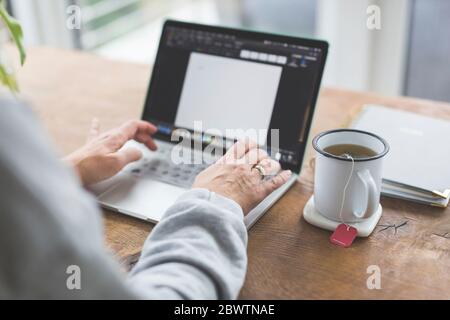 Donna che lavora su un computer portatile, con una tazza di tè sul lato Foto Stock