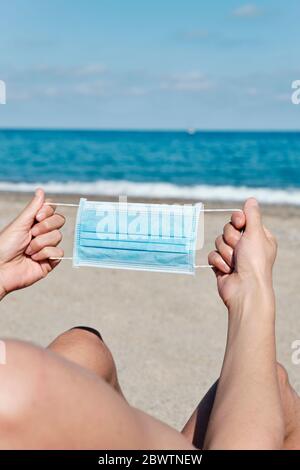 primo piano di un giovane caucasico con una maschera chirurgica blu nelle mani sulla spiaggia Foto Stock