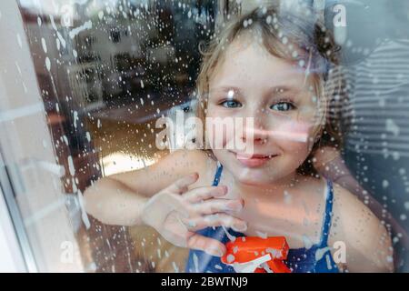 Ritratto di bionda sorridente con flacone spray che guarda attraverso la finestra bagnata Foto Stock