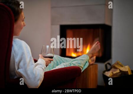 Donna seduta su poltrona con bicchiere di vino rosso che si rilassa davanti al camino Foto Stock