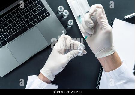 Immagine ritagliata della siringa per iniezione manuale del medico nel flaconcino sul computer portatile alla scrivania del laboratorio Foto Stock