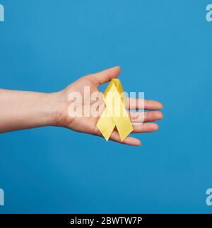 mano femminile tenere un nastro giallo sotto forma di un loop su sfondo blu, simbolo della lotta contro il tumore del gatto, problema di suicidi e thei Foto Stock