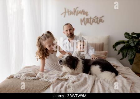 Bambina stroking il suo cane sul letto mentre padre e brothersitting in background Foto Stock