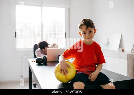 Mamma che lavora a casa con i suoi figli, figlio seduto sul tavolo con la palla da calcio Foto Stock