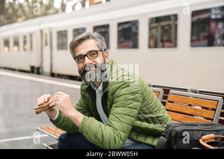 Uomo seduto su una panchina alla stazione ferroviaria che mangia un panino Foto Stock