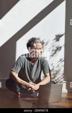 Uomo maturo seduto sul pavimento con un computer portatile che guarda a distanza Foto Stock