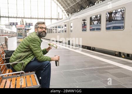 Uomo seduto su una panchina alla stazione ferroviaria con sandwich e bevanda calda Foto Stock