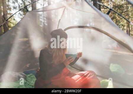 Giovane donna seduta in tenda, mangiando la colazione nella foresta Foto Stock