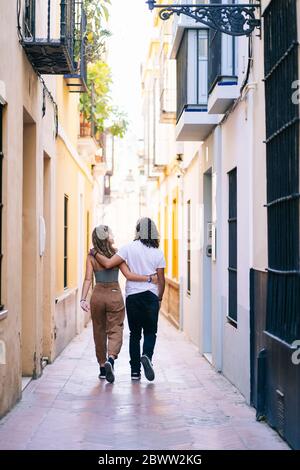 Vista posteriore di una giovane coppia che cammina con le braccia intorno su una strada stretta a Santa Cruz, Siviglia, Spagna Foto Stock