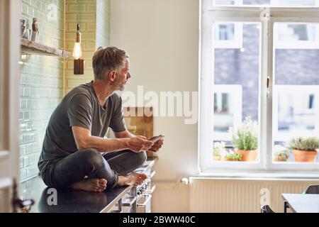 Uomo maturo con telefono cellulare seduto sul banco da cucina a casa Foto Stock