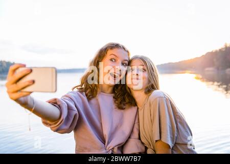 Amici felici che fanno i volti mentre si prendono selfie su smartphone contro lago Foto Stock