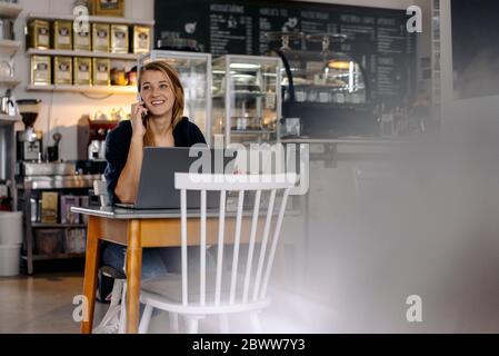 Giovane donna sorridente che usa smartphone e computer portatile in un bar Foto Stock