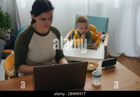 Donna che lavora su un computer portatile a casa mentre suo figlio guarda i film sul telefono cellulare Foto Stock