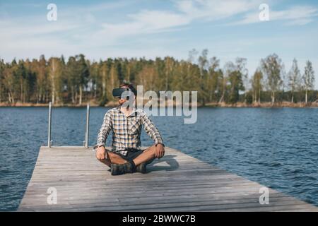 Giovane seduto sul molo, indossando occhiali VR Foto Stock