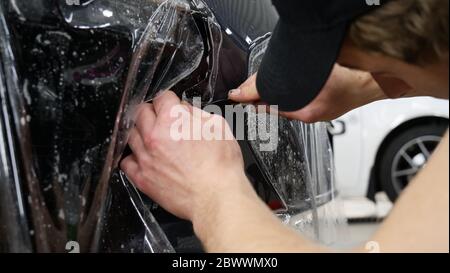 Primo piano sul processo di installazione del PPF su un faro anteriore e su un cofano. PPF è una pellicola di protezione della vernice che protegge la vernice da graffi e scheggiature. Foto Stock