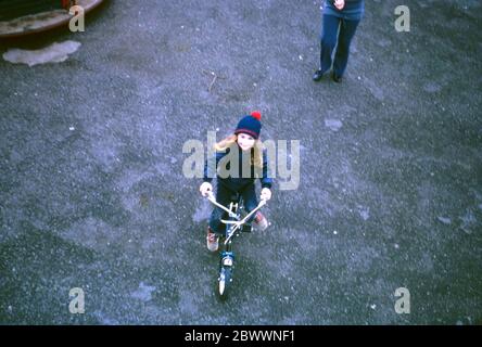 Una ragazza di 7 anni che indossa jeans blu, un cappello di lana blu con una bobina rossa e una giacca blu, che cavalca la sua bici Raleigh Tomahawk intorno a una rotonda parco giochi, Hatch End, Middlesex, Regno Unito Foto Stock