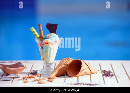 Panorama di vetro con gelato arcobaleno e sciroppo colorato su sfondo bokeh sfocato con spazio copia. Cialda di coni di cioccolato su tavolo bianco del blu Foto Stock