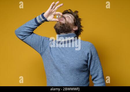 Shaggy, uomo bearded in un maglione beve alcool forte da un wineglass, su uno sfondo giallo. Concetto di alcolismo. Foto Stock