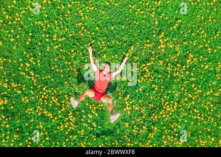 Felice uomo sorridente felice sorridente e ridente sdraiato in abiti rosso e arancio luminoso sull'erba con dente di leone giallo in giornata di sole, vista aerea dall'alto Foto Stock