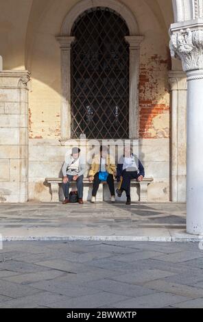 Tre turisti stanchi si rilassano su una panchina sotto e arco al Palazzo Ducale di Venezia Foto Stock