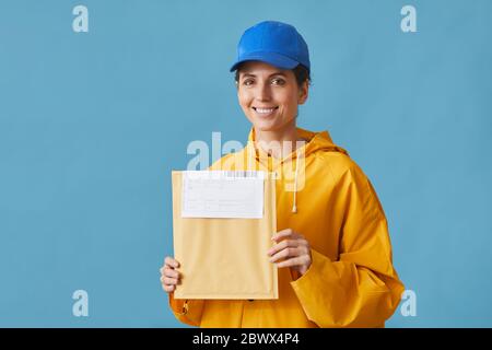 Ritratto di giovane postdonna in uniforme che tiene lettera e sorridendo alla macchina fotografica contro lo sfondo blu Foto Stock