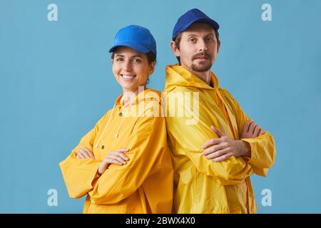 Ritratto di giovane coppia in uniforme che lavora in servizio di consegna in piedi con le braccia incrociate e sorridenti alla fotocamera contro lo sfondo blu Foto Stock