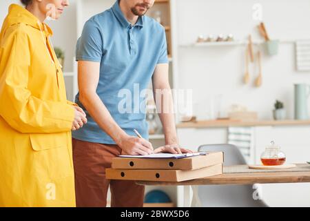 Primo piano di giovane uomo che firma il modulo per la pizza mentre donna consegna la pizza a casa Foto Stock
