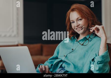 Positivo buona donna giovane ragazza libera funziona a distanza su computer portatile, collegato a internet wireless gratuito a casa, tiene gli occhiali, vestito Foto Stock