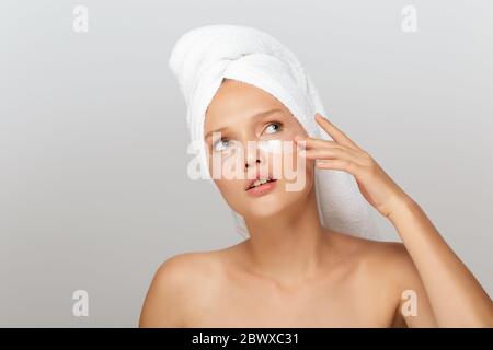 Ritratto di giovane donna con asciugamano bianco sulla testa senza trucco con macchie trasparenti sotto gli occhi guardando pensieroso su sfondo grigio Foto Stock