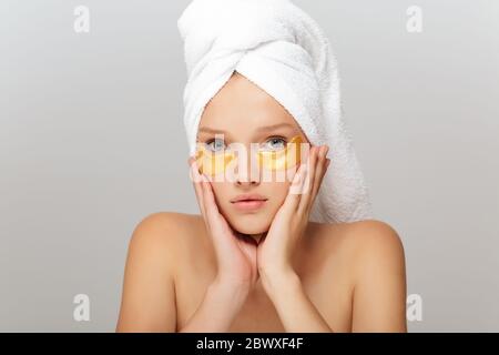 Ritratto di giovane donna con asciugamano bianco sulla testa senza trucco con macchie gialle sotto gli occhi guardando pensieroso in macchina fotografica su sfondo grigio Foto Stock