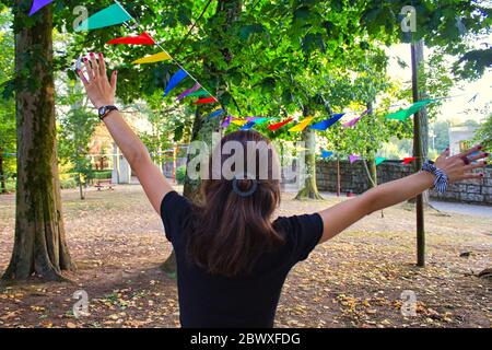 Giovane donna con le braccia in su divertirsi in una festa in un parco con decorazioni bandiere di colori diversi Foto Stock