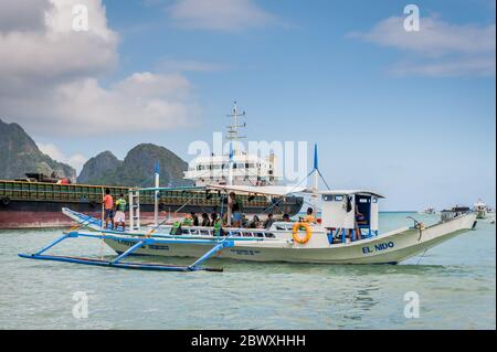 Una barca di pescatori dalla coda lunga carica di rifornimenti e turisti per fare una gita di un giorno con una enorme petroliera alle spalle dell'isola di El Nido, Filippine. Foto Stock