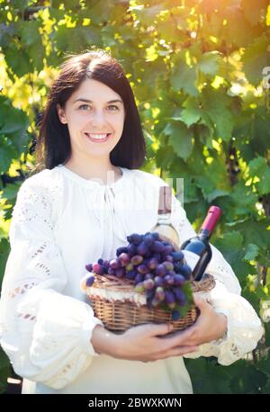 Giovane donna con un cesto di uve e bottiglie di vino in vigna. Foto Stock