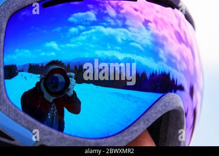 Fotografo e fotocamera riflessa in occhiali in inverno con neve e montagne. Auto foto all'aperto vicino alla foresta, sulla pista da sci e snowboard Foto Stock