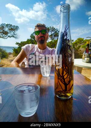 uomo in vacanza estiva sulla spiaggia seduto ad un tavolo da ristorante con una bottiglia di bicchiere di liquore alle erbe hierba a ibiza Foto Stock