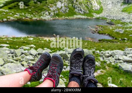 Giovane coppia innamorata mostrando i loro stivaletti stanchi dopo aver scalato gli alti sentieri con il bellissimo lago glaciale sullo sfondo Foto Stock