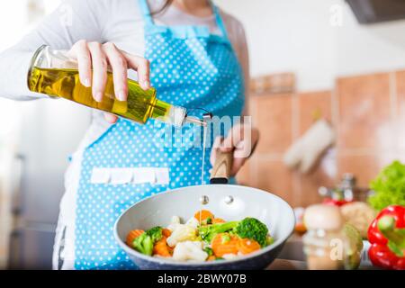 Donna che versa olio d'oliva sulle verdure in padella a casa. Concetto di pasto sano. Foto Stock