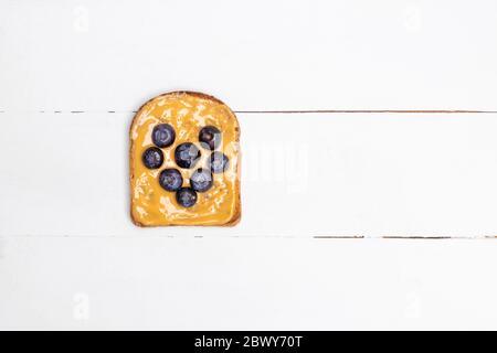 Sano tostato vegano con burro di arachidi e mirtillo a forma di cuore su tavolo di legno bianco. Vista dall'alto con spazio per la copia Foto Stock